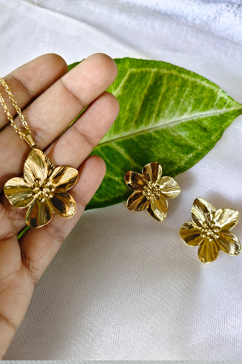 A gold-tone flower pendant necklace and matching stud earrings displayed on a white fabric surface, with a hand holding the pendant and a green leaf prop behind it.