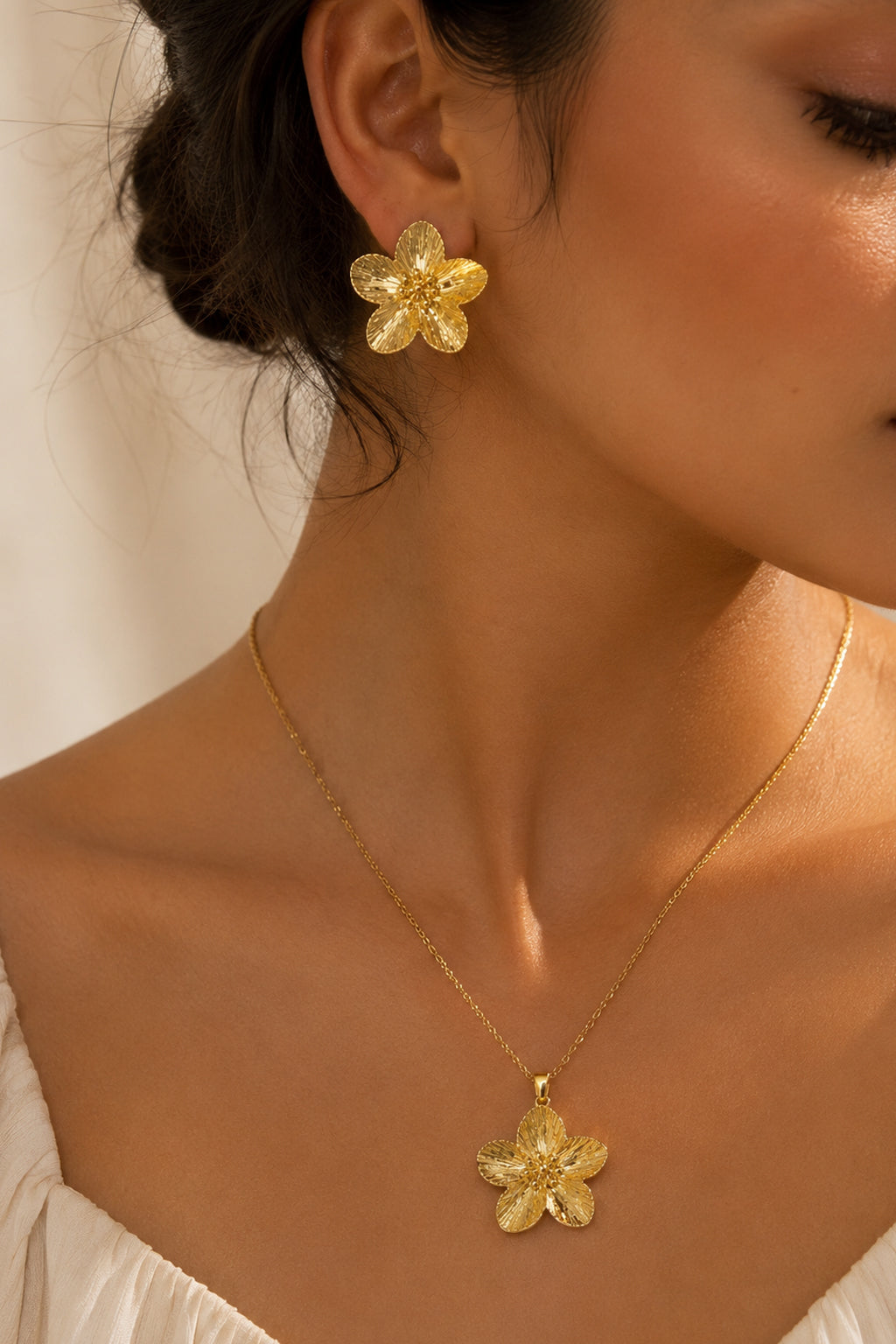 A close-up lifestyle shot of a model wearing a matching gold flower-shaped stud earring and necklace with a textured gold flower pendant.