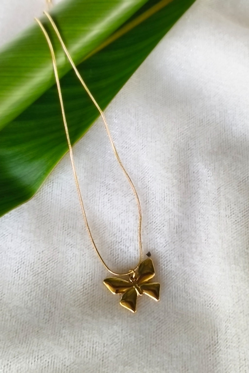 A delicate gold necklace with a small bow-shaped pendant, resting on a textured white fabric surface next to a green leaf.