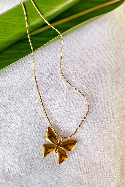 A gold necklace with a textured, faceted butterfly pendant, displayed on a light beige fabric background with a green leaf prop.