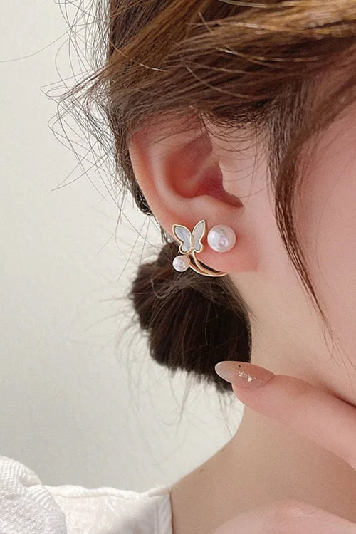A close-up of a person's ear wearing a gold butterfly-shaped stud earring with white faux pearls, set against a soft off-white background.